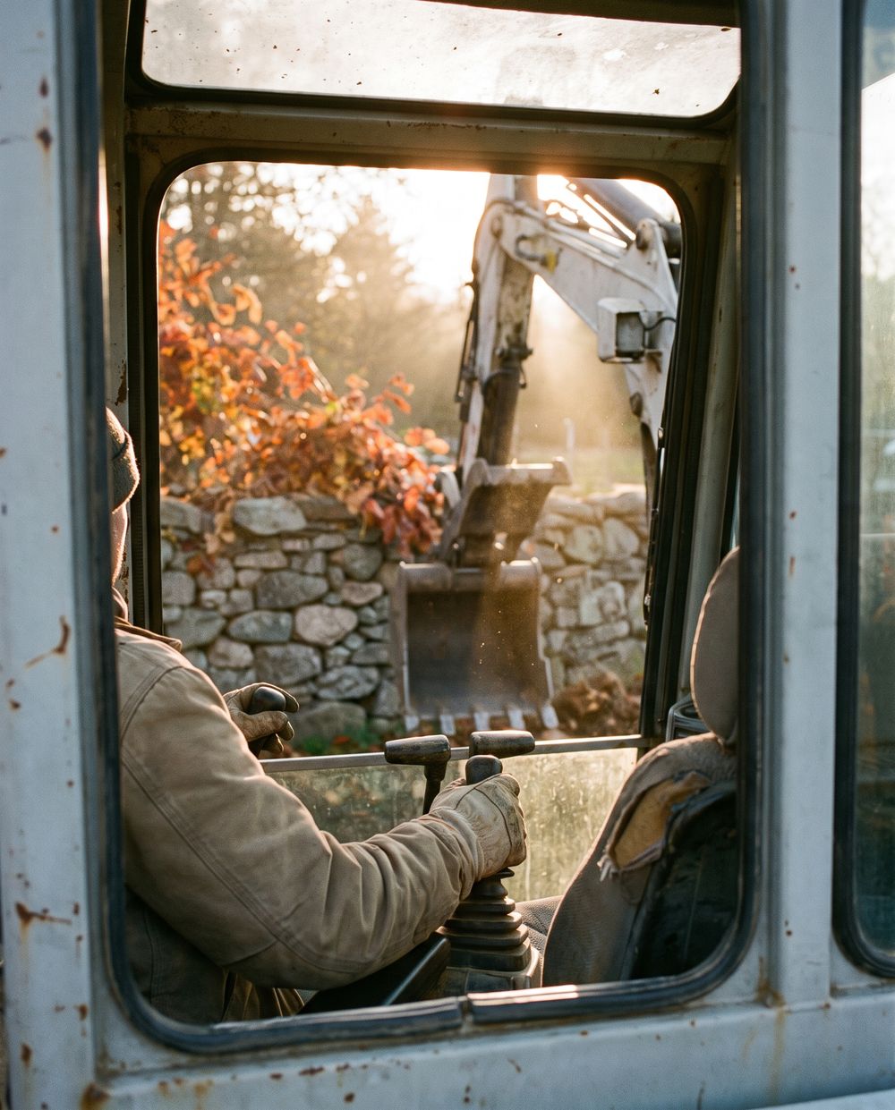 View from an excavator cab: gloved hands on the controls, a fieldstone wall and fall foliage through the glass